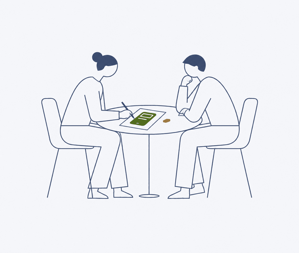 Two people at a small table, one sketching a single product screen on paper while the other listens